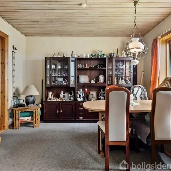 En spisestue med et træbord omgivet af polstrede stole. Skænk med glaslåger står bagved fyldt med porcelæn. Vinduet til højre slipper lys ind, og en loftlampe hænger over bordet.