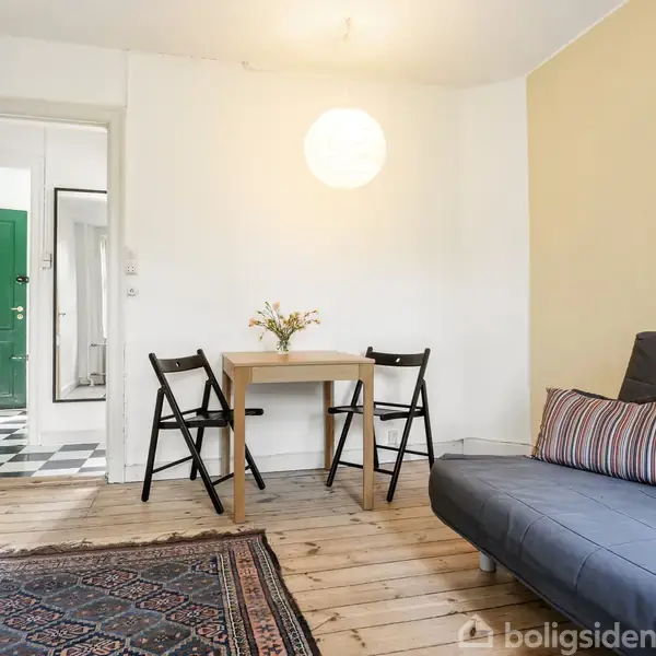 Et rum med en bord og to stole står foran en hvid væg. En grå sofa med puder er til højre, og en farverig tæppe ligger på trægulvet.