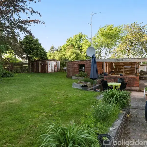 En græsplæne med borde og stole under en parasol ved siden af en murstensbygning i en have med træer og buske rundt omkring.