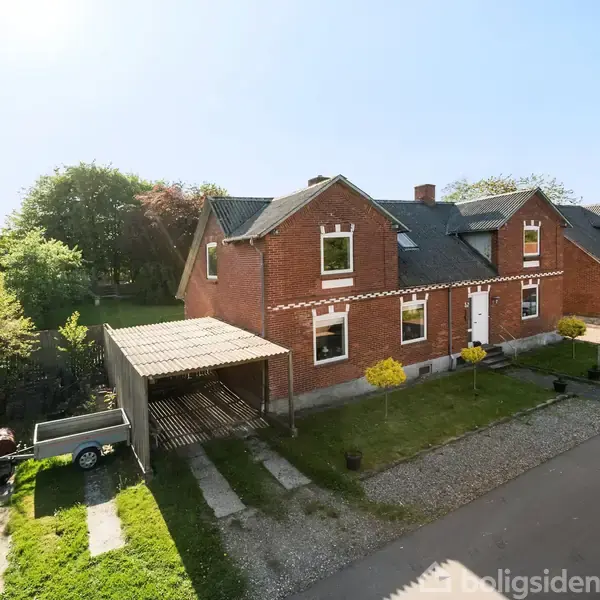 Rødt murstenshus står stille med to etager og hvide vinduer. Tilknyttet carport med trailer. Omgivet af græsplæne og træer på solsiden af en stille vej.