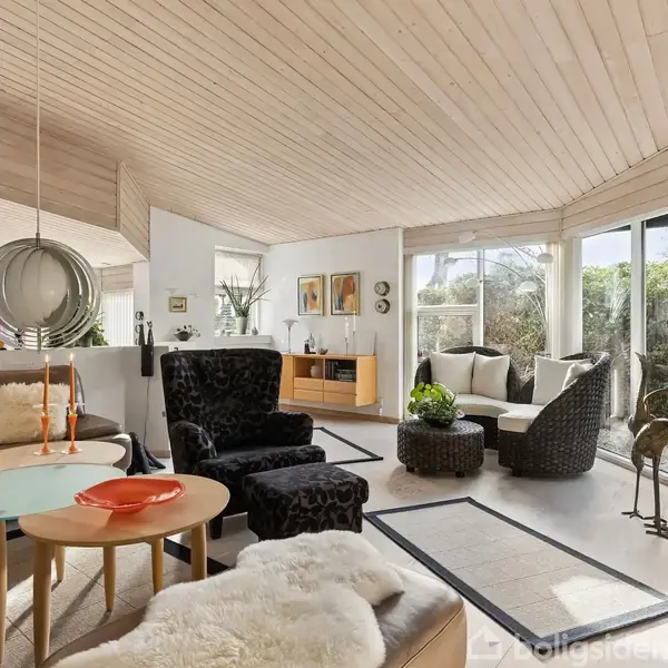 Stue med lædersofa, sofabord og lænestol indrettet i moderne stil. Store vinduer lukker naturligt lys ind. Træloft og grønne planter skaber en lys og indbydende atmosfære.