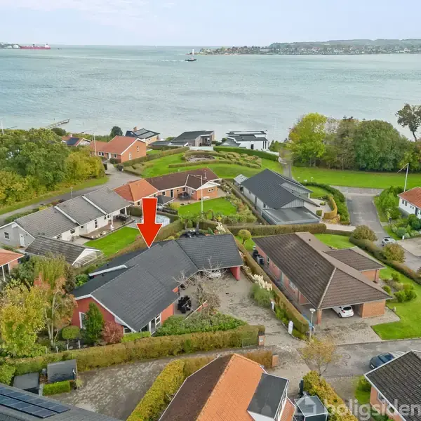 Rød pil peger på en sorttagshus i et boligkvarter. Husene ligger tæt, omgivet af grønne haver. Baggrunden viser en bred udsigt over en fjord med skibe og modsat kystlinje.