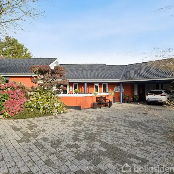 Hus med orange facade og mørkt tag, omgivet af planter, står stille på en stenbelagt gårdsplads med en bil parkeret i carporten til højre.