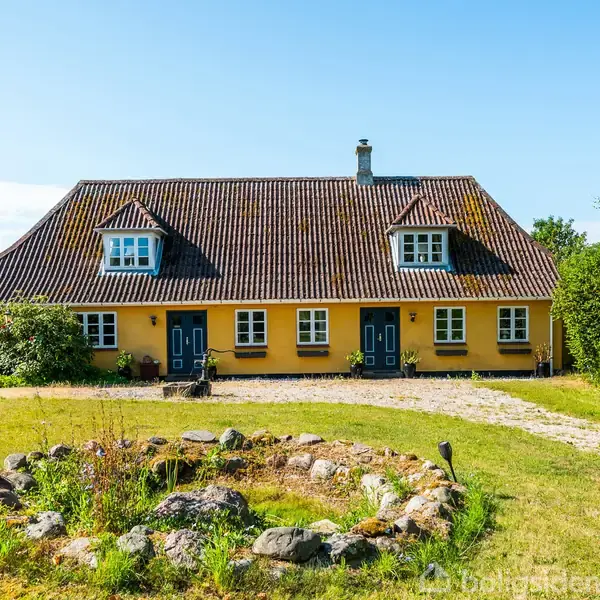 Gul bindingsværkhus står i et grønt landskab. Huset har to etager med to kviste på taget, omgivet af græs og buske. En stenbelagt sti leder til hoveddøren.