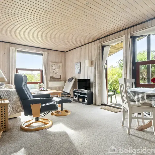 Stue med lænestol og lampe, placeret ved siden af et sofabord. Et spisebord med stole står ved vinduet, der åbner ud til en terrasse med udsigt til haven.