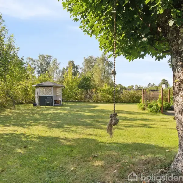 Et reb hænger fra et træ med en forgrening. Rebgyngen svajer forsigtigt i en stor, grøn have med græsplæne, buske og et lille lysthus i baggrunden.