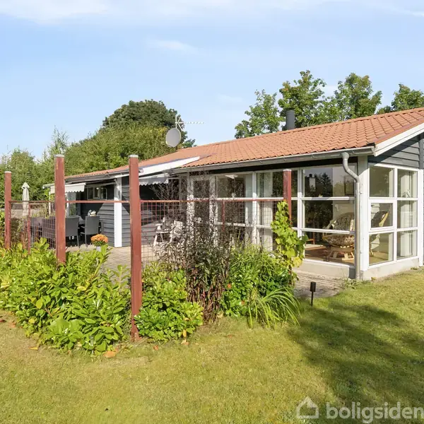 Et sort træhus med rødt tag står i en frodig have, omgivet af tæt vegetation og grøn plæne. Huset har store vinduer og en tilhørende terrasse.