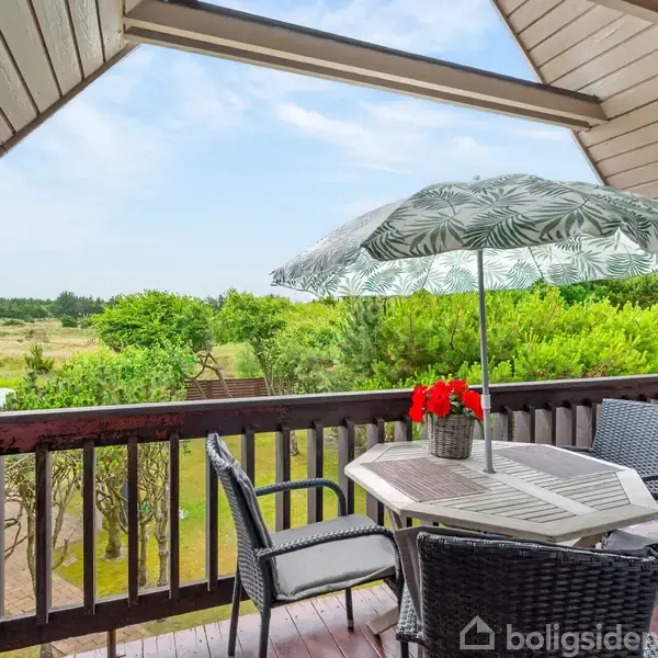 Terrassebord med stole omkring er dækket af en parasol. Røde blomster i midten. Omgivet af grønne buske og træer, med udsigt til natur og himmel.