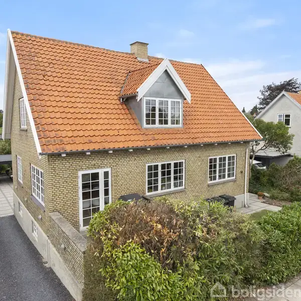 Et to-etagers murstenshus med rødt tegltag står i en forstad. Huset har en stor gavlvindue og er omgivet af velplejede hække og en flisebelagt indkørsel.