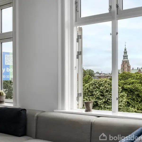 En stue med en lang sofa langs vinduerne. Udsigt over trætoppe og tårne i byen udenfor. På vindueskarmen står små planter og puder ligger på sofaen.