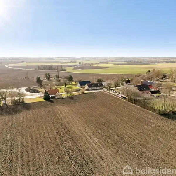Landskab med åbne marker strækker sig ud. En lille samling huse ligger tæt ved skov. Sollys belyser markerne, og en vej snor sig gennem landskabet i baggrunden.