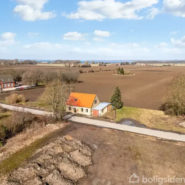 Hus med orange tag og hvide vægge står stille ved vej, omgivet af åbne marker og få træer i landligt område under en blå himmel med skyer.