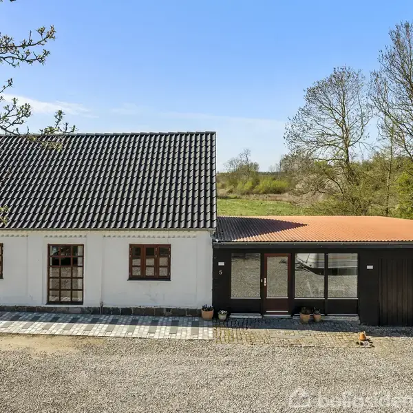 Hvidt hus med sort tag og tilstødende carport ses i en landlig omgivelser. Der er en grusvej foran og træer i baggrunden.