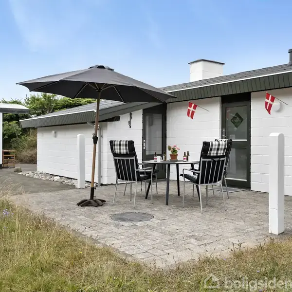 Et hvidt sommerhus med sort tag har en terrasse med et bord og fire stole under en parasol. Danske flag er hængt op ved døren. Græsset omkring er grønt.