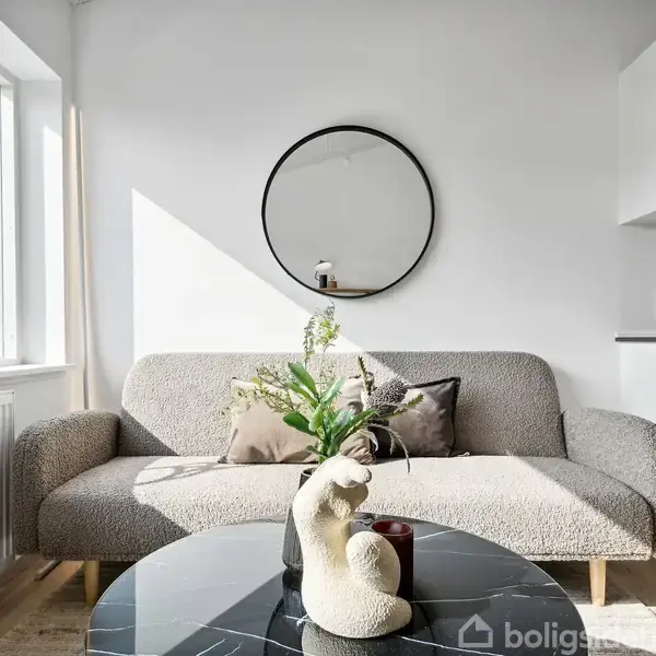Sofa står i en lys stue ved siden af et vindue. Et spejl hænger på væggen, og en skulptur samt planter står på et rundt sofabord foran sofaen.