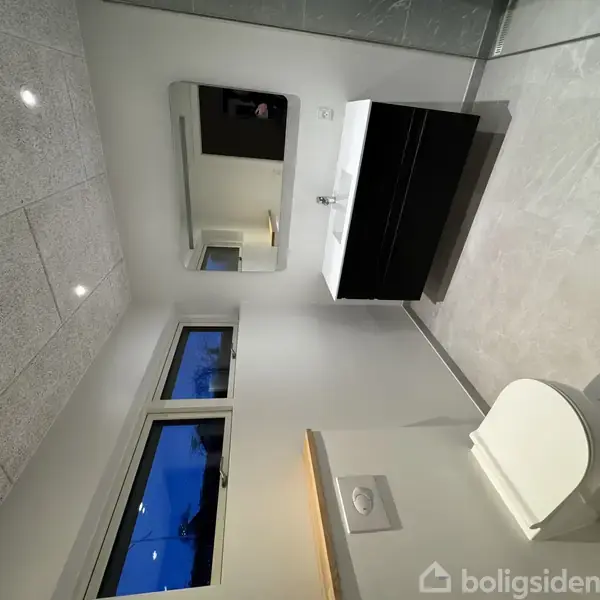 Toilet placeret ved siden af vask med spejl på væggen; badeværelse med lyse vægge, flisegulv og vindue.