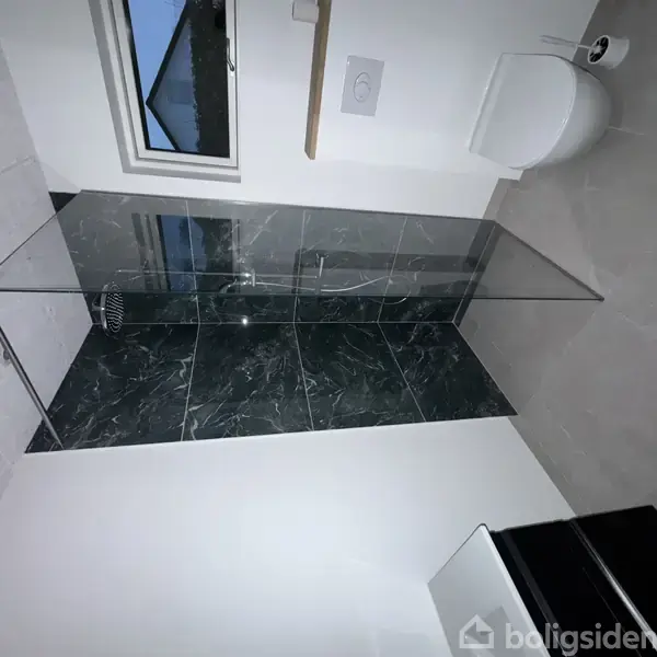 Brusekabine med klart glas og mørke fliser, placeret i et moderne badeværelse med hvidt toilet, lyse vægge og gulvfliser, samt et vindue i baggrunden.