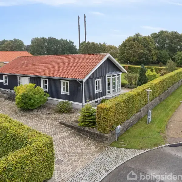 En sortmalet étplanshus med rødt tag står stille på en brolagt indkørsel, omgivet af grønne hække og græsplæne. Baggrund med skov og en sti langs haven.