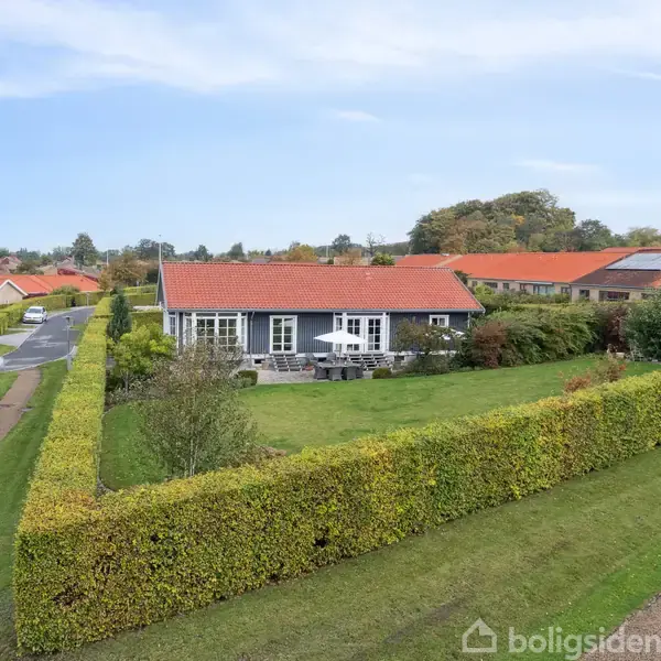 Hus med rødt tag og hvide vinduer står omgivet af velplejede grønne hække. Græsplæne og gangsti ses i forgrunden, omgivet af flere huse med lignende røde tage.