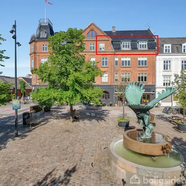 Bygning med rød murstensfacade har en markeret lejlighed. I forgrunden ses en skulptur med en fugl i en springvand, omgivet af træer på en brostensbelagt plads.