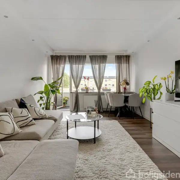 En lys stue med en beige sofa fyldt med puder, en rund sofabord, og et stort TV på et hvidt skab. Store vinduer med gardiner og planter i baggrunden.