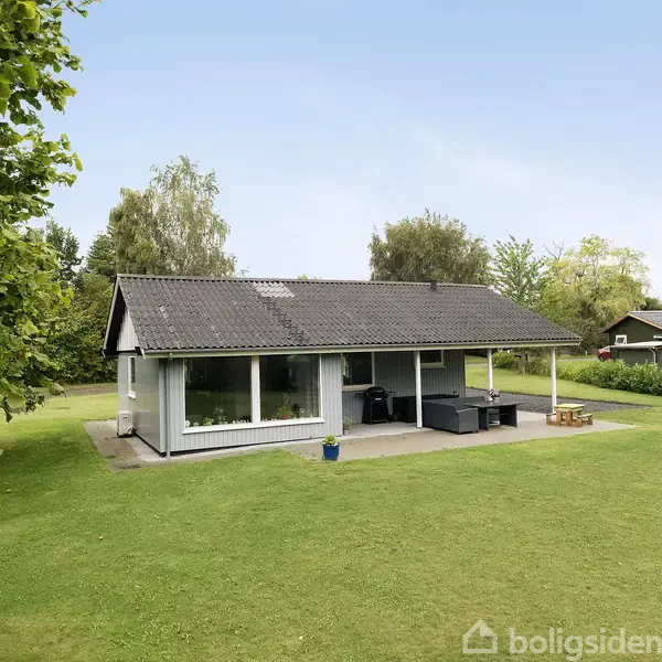 Et enkelt hus står på en grøn græsplæne, omgivet af træer og buske. Terrasse møbler er placeret foran huset, der har store vinduer og et mørkt tag.