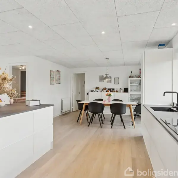 Et moderne køkken med hvide skabe og en keramisk kogeplade. Ved siden af er der et spisebord med fire stole. Rummet er lyst med store vinduer og en åben planløsning.