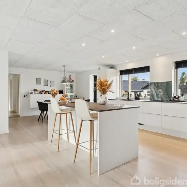 Et moderne køkken med en ø, barstole og blomster i vase, omgivet af lyse skabe og vinduer; åbner op til en spisestue og en dør til et soveværelse.