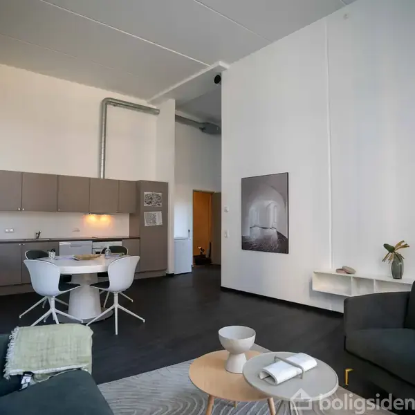 Stue med to sofaer og sofabord, nær åben køkken med spisebord og stole. Minimalistisk indretning med en dekorativ plakat på væggen. Højt til loftet og mørkt gulv.