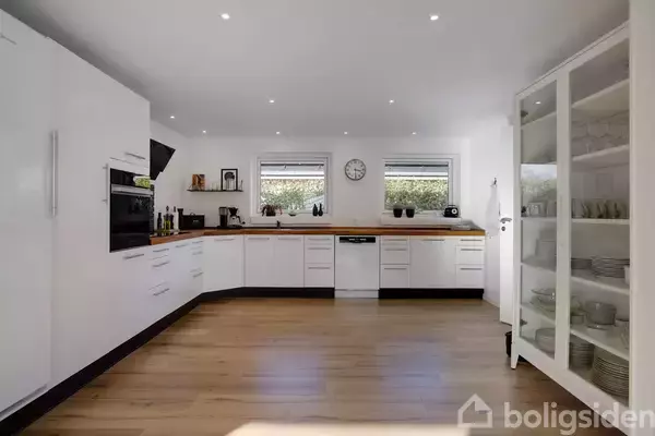 Et moderne køkken med hvide skabe og trægulv. Køkkenet har en indbygget ovn, opvaskemaskine, og to vinduer. Der er en stor skænk med glaslåger fyldt med service i højre side.