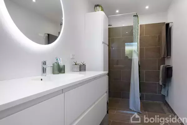 Badeværelsesskab med hvid vask, spejl med lys omkring, grøn plante og tandkrus på bordpladen. Bruseniche med brun flisevæg i baggrunden. Rummet har belysning i loftet.