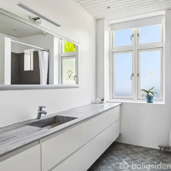 Moderne badeværelse med langt, hvidt skabsmodul og integreret vask. Over vasken hænger et stort spejl, og i baggrunden ses et vindue med plante. Rummet har lyse farver.