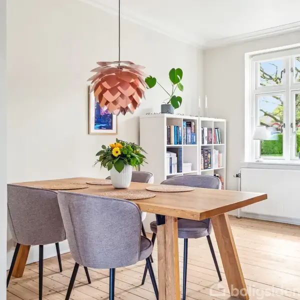 Træbord med grå stole står i spisestue, pyntet med blomster i vase. Over bordet hænger en moderne lampe. Bagerst er en bogreol ved siden af et vindue med udsigt til haven.