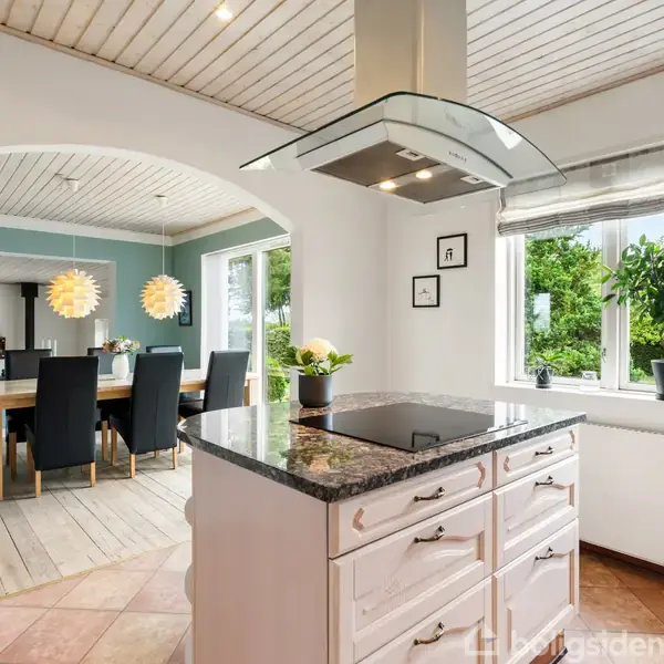 Køkkenbord med emhætte over står i forgrunden, omgivet af hvidmalede vægge. Til venstre er et spiseområde med spisebord, stole og to dekorative lamper. Store vinduer giver udsigt til en grøn have.