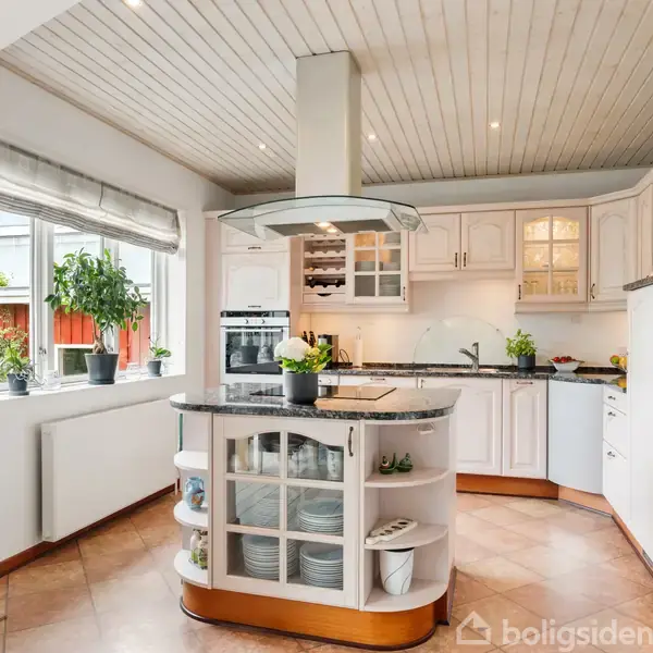 Et lyst køkken med en central ø, der har hylder og opbevaring. Køkkenet har hvide skabe, integrerede apparater og vinduer, der giver naturligt lys. Grønne planter pynter rummet.