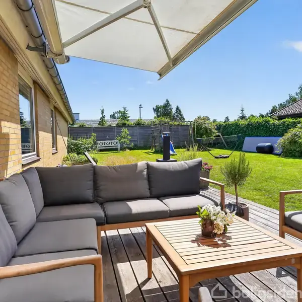 Grå sofaer står omkring et træbord på en terrasse. En markise giver skygge, og i baggrunden ses en have med græs og buske.