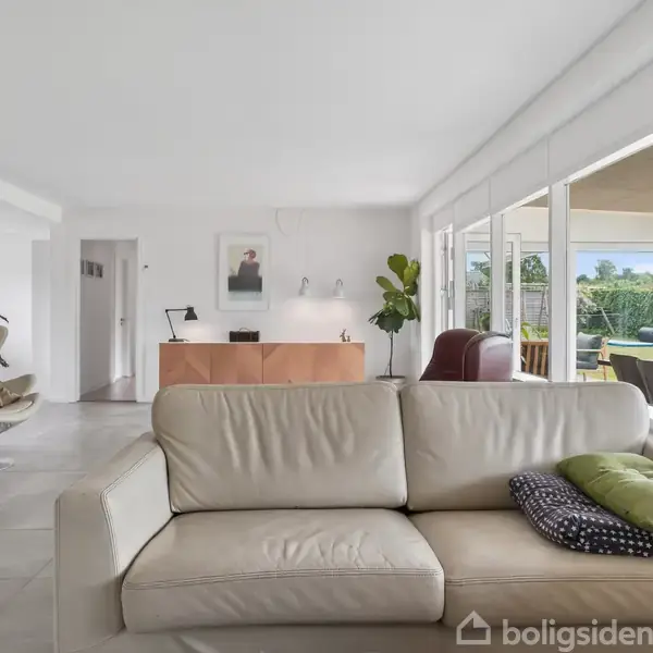 Beige sofa står i en lys stue med store vinduer, udsigt til en terrasse. Et billed hænger på væggen, planter og moderne møbler supplerer rummet.