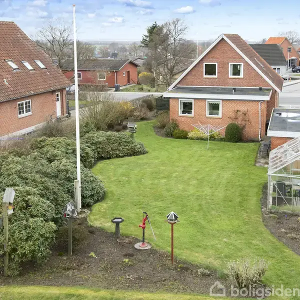Et rødt murstenshus med græsplæne foran. Til højre står et drivhus. Buske omkranser haven. Der er en flagstang i midten, og stier findes i nabolaget.