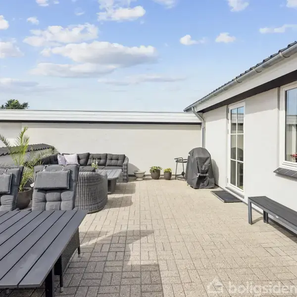 Terrasse med grå fliser fyldt med havemøbler og planter. Sofaer og et bord står under en blå himmel ved siden af et hvidt hus med store vinduer.