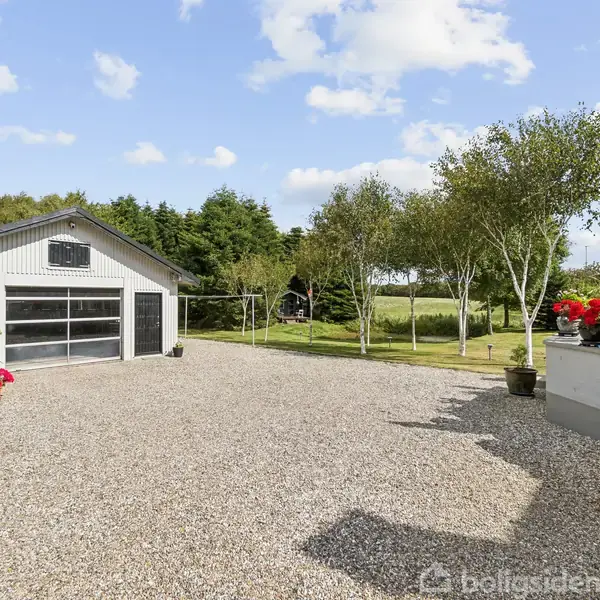 Grusgårdsplads med blomsterkrukker og garage i baggrunden. Sollys skinner på stien omgivet af træer og frodig natur. Stole og paraplyer står på terrassen til højre.