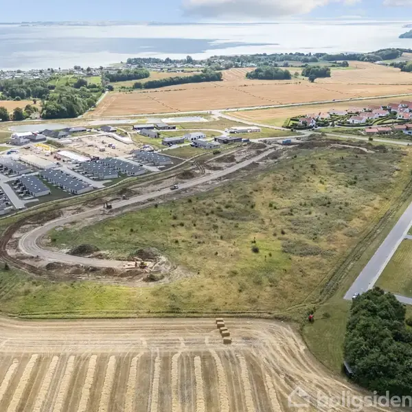 En stor åben mark med en snoet grusvej i centrum omgivet af byggepladser, nybyggeri og et etableret boligområde. I baggrunden ses marker og fjorden i horisonten.