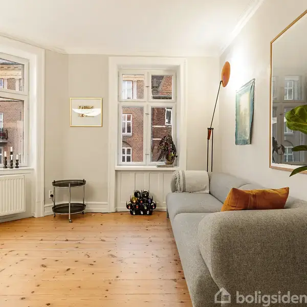 En grå sofa med orange pude står i en lys stue med trægulv; store vinduer giver udsigt til en murstensbygning. Der er en plante til højre.