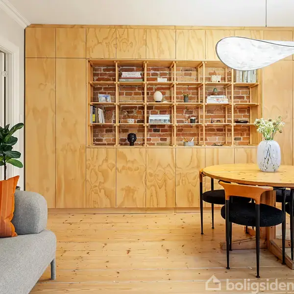 En trævæg med indbygget reol fylder rummet, med bøger og dekorationer. Et rundt bord med sorte stole står foran, mens en grå sofa med orange pude er på venstre side.