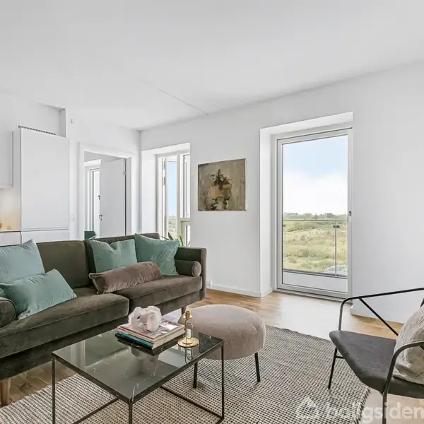 En moderne stue med sort sofa og grønne puder står ved et glasbord. Ved siden af er en lænestol og gulvlampe. Store vinduer viser en udsigt over naturen udenfor.