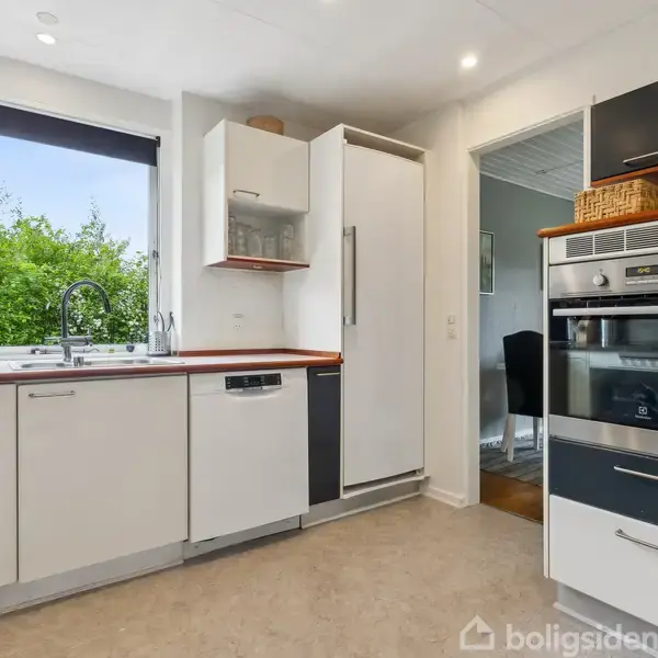 Moderne køkken med sorte og hvide skabe. En opvaskemaskine og en ovn er indbygget. Store vinduer giver udsigt til en frodig have. En dør fører til et tilstødende rum.