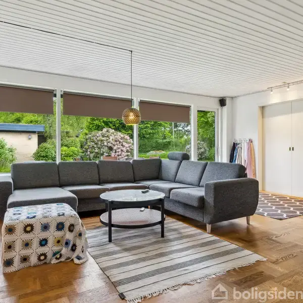 Grå sofa står stille i stue med trægulv. Store vinduer med rullegardiner åbner mod en grøn have. To tæpper ligger på gulvet ved sofabordet.