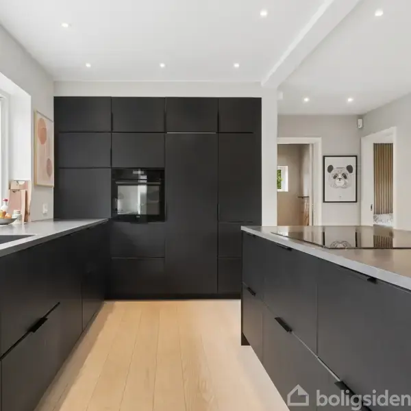 Sort køkken med moderne design; centralelement er en indbygget ovn i væggen; køkkenø med sort overflade, omgivet af store vinduer, der giver naturligt lys.