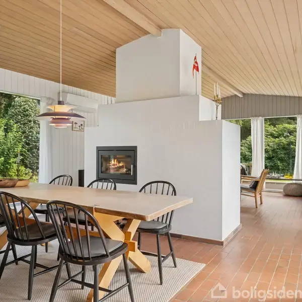Et spisebord med sorte stole står i et lyst rum med store vinduer. En pejs er indbygget i en hvid væg, og der er udsigt til en grøn have.