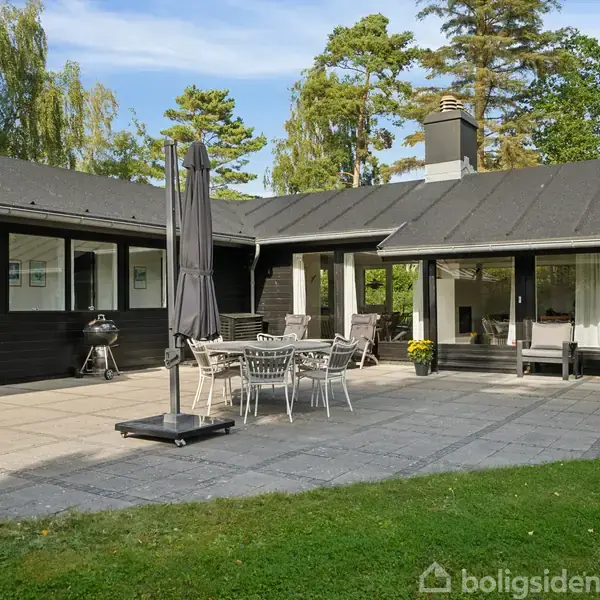 En terrasse med havemøbler og en grill står foran et sortmalet træhus med store vinduer. Haven er grøn med træer i baggrunden.