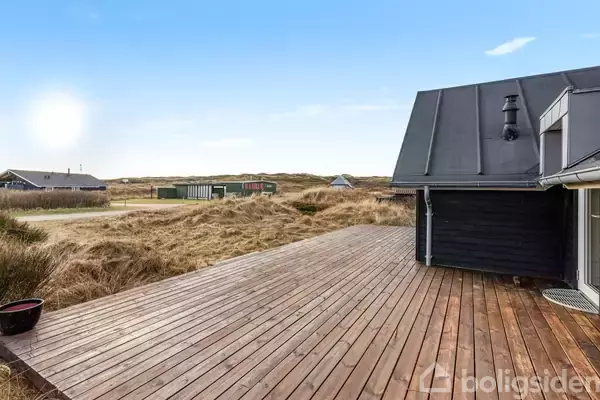 En træterrasse strækker sig fra et sort, moderne sommerhus med græsland omkring og flere huse i horisonten under en blå himmel.